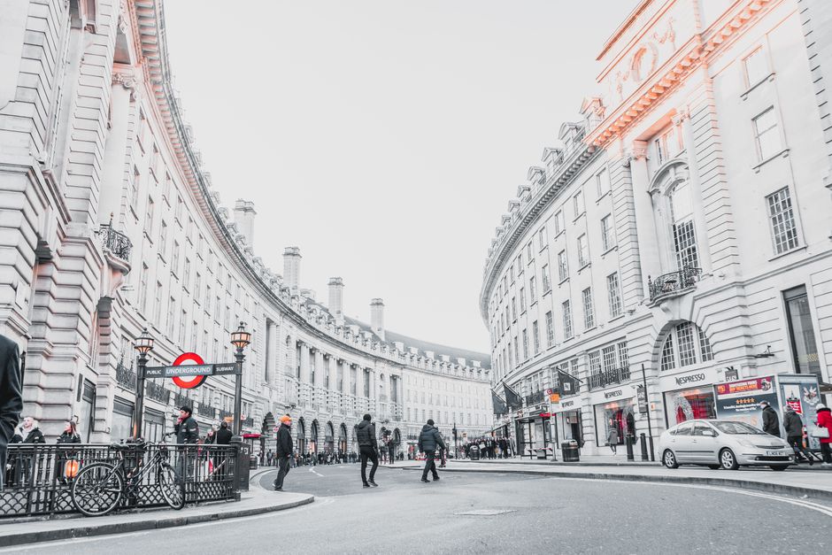 Nighttime view of a London underground entrance with a circular red and blue 'Underground' sign, illuminated lamp posts, and historic buildings with detailed facades in the background. The scene featu