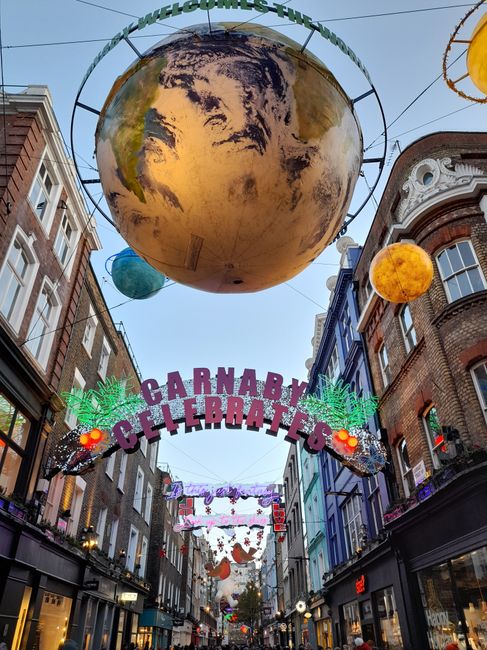 Nighttime scene on Carnaby Street in Soho, W1D, London, showing a crowded pedestrian area with people walking along a wet cobblestone surface illuminated by festive string lights and colorful shop fac